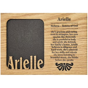 May include: A wooden picture frame with a black mat and a cutout of the name "Arielle" in the bottom left corner. The frame has a quote about the name Arielle, which is of Hebrew origin and means "lioness of God".
