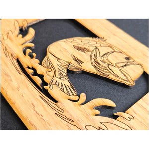 May include: A light brown wooden cutout featuring a laser-engraved design of a fish jumping out of water. The fish is depicted in profile with detailed line work for its scales and fins, set against a dark background within a rectangular frame.
