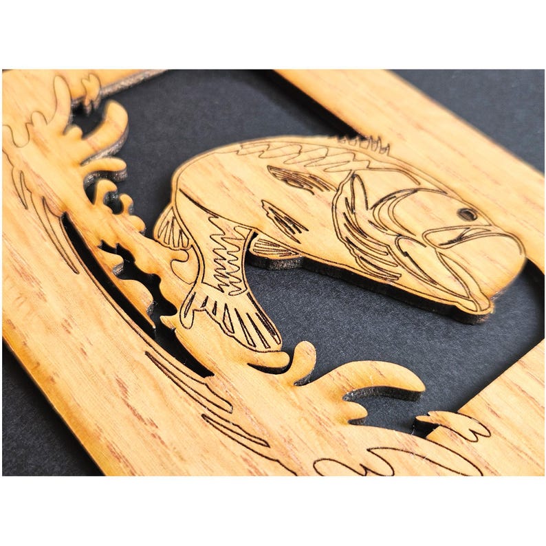 May include: A light brown wooden cutout featuring a laser-engraved design of a fish jumping out of water. The fish is depicted in profile with detailed line work for its scales and fins, set against a dark background within a rectangular frame.