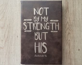 May include: A brown leather-bound journal with the words "NOT BY MY STRENGTH BUT HIS" in white lettering. The text "Zechariah 4:6" is printed in smaller white lettering below the main text.