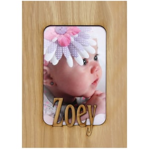 May include: A wooden photo frame with a cutout for a picture and a personalized name. The frame is light brown and has a natural wood grain. The picture shows a baby girl wearing a flower headband. The name "Zoey" is cut out of the wood below the picture.
