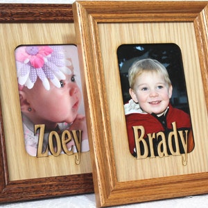 May include: Two wooden picture frames with a light brown wood finish. The frames are side by side. The left frame has a photo of a baby girl with a pink flower headband. The name "Zoey" is cut out of the wood below the photo. The right frame has a photo of a young boy. The name "Brady" is cut out of the wood below the photo.