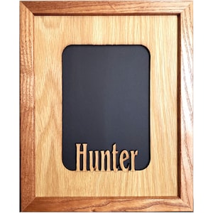 May include: A rectangular wooden picture frame with a light brown wood grain finish. The frame features a central vertical rectangular opening with rounded corners, backed by a solid black panel. The name Hunter is cut out in wood.