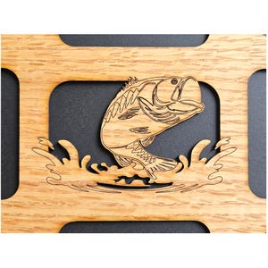 May include: A light brown wooden cutout featuring a laser-engraved largemouth bass jumping out of water. The fish is depicted in a dynamic pose with detailed scales and fins, set against a dark background within a rectangular wooden frame.