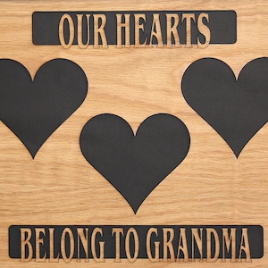 Puede incluir: Señal de madera con el texto "Our Hearts Belong to Grandma" y tres recortes de corazones negros.