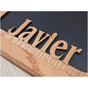 May include: A wooden sign featuring the name Javier in cut-out letters. The letters are crafted from light-colored wood with a visible grain, mounted onto a dark, matte black rectangular background. The sign has a thick wooden frame border.