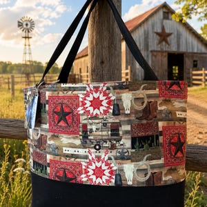 May include: A tote bag with a Western theme, featuring a black base and black straps. The upper portion displays a patchwork design with red stars, bandanas, cowboy boots, and steer skulls. The background includes a barn, windmill, and a wooden fence.