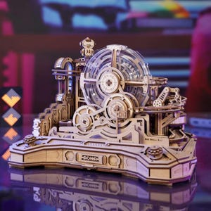 Puede incluir: Maqueta mecánica de madera con engranajes, una esfera transparente y la palabra "ROKR" en la base. El modelo presenta detalles intrincados, incluyendo escaleras, rampas y una pequeña figura en la parte superior. El diseño sugiere una construcción interactiva compleja.