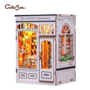 May include: A detailed miniature model of a bookstore, labelled "The Scholar's Bookstore". The white building has brickwork and a lit interior, revealing books and decorative items. An open window is also visible, adding to the model's charm.