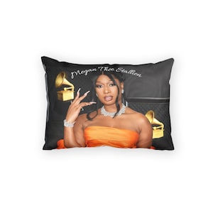 May include: Rectangular pillow featuring a portrait of Megan Thee Stallion in an orange strapless top, with a diamond necklace and bracelet. The pillow has a black background with two gold gramophone statues and the text "Megan Thee Stallion".