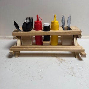 Pode incluir: Uma pequena mesa de piquenique de madeira contém utensílios e condimentos. Colheres, garfos e facas pretas são colocados à esquerda e à direita. Frascos vermelhos e amarelos, e recipientes pretos e brancos estão no centro.