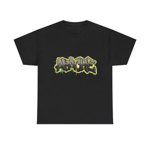 Puede incluir: Camiseta negra con la palabra "APACHE" en estilo grafiti. El texto es gris con contorno amarillo y detalles verdes, creando una estética audaz y urbana. Estilo urbano.