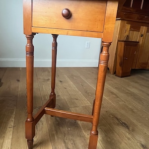 May include: A small, wooden side table with a single drawer and a round knob. The table has four slender, turned legs and a cross-brace for stability. The wood has a warm, reddish-brown finish.