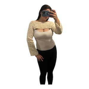 May include: A cream-colored, long-sleeved crochet shrug featuring an open-knit, grid-like pattern. The garment has a wide, boat-style neckline and covers the arms and shoulders, worn over a solid, light-colored tank top and black leggings.