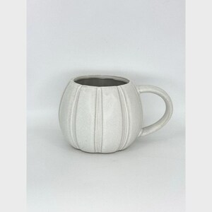 May include: A round, off-white ceramic mug with a textured, ribbed surface that mimics the shape of a pumpkin. The mug features a smooth, curved handle on the right side and a wide, open top with a light gray interior finish.