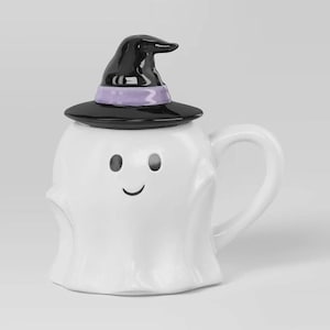 May include: A white ceramic mug shaped like a ghost with a smiling face, featuring a removable black witch hat lid with a lavender band. The mug has a curved handle on the right side and a smooth, glossy finish throughout the entire piece.
