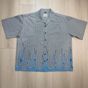 Op de afbeelding: Een overhemd met korte mouwen en knoopsluiting, voorzien van een grijs traanplaatpatroon over het hele kledingstuk. De onderzoom is versierd met een reeks lichtblauwe en donkerblauwe vlammen die over de voorkant van het overhemd lopen.