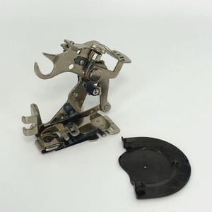May include: A silver-colored sewing machine attachment with multiple parts, including a curved arm and a small, black, semi-circular piece. The attachment is designed for sewing and is likely used for a specific stitch or technique.