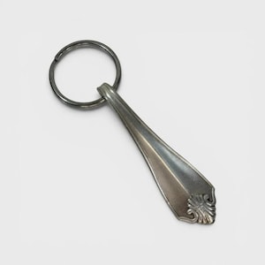 Puede incluir: Un llavero de metal plateado que presenta una anilla circular unida a un segmento de mango cónico de una pieza de cubertería. El mango tiene una superficie lisa y pulida con un patrón decorativo en relieve en forma de abanico en el extremo.