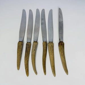Puede incluir: Un juego de seis cuchillos de mesa con hojas de metal plateado y mangos texturizados de color marrón claro que se asemejan al material de asta natural. Los cuchillos están dispuestos verticalmente en una fila paralela y uniforme.