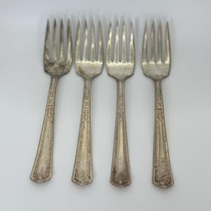 May include: Four antique silver forks with ornate, detailed handles and tines. The forks are arranged in a row against a white background. The silverware shows signs of age and use, with some tarnishing visible on the metal.