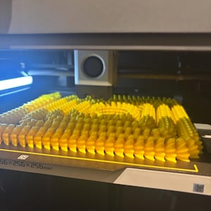 Puede incluir: Una impresora 3D muestra múltiples objetos cilíndricos amarillos y puntiagudos en proceso de impresión. Los objetos están dispuestos en filas sobre una superficie negra. La impresora tiene un marco gris y negro.