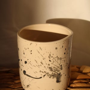 Peut inclure: Un gobelet en céramique de couleur crème avec un motif de peinture noire mouchetée. La tasse cylindrique a un bord légèrement incurvé et convient aux boissons. La tasse mesure environ 10 cm de haut.
