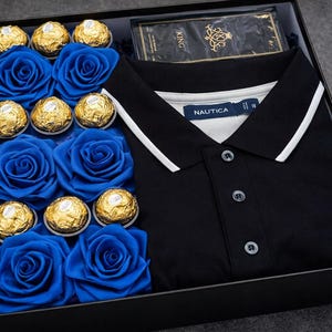 Peut inclure: Un coffret cadeau noir contenant un polo noir avec un col blanc, des roses bleues et des chocolats enrobés d'or. La chemise porte la marque "NAUTICA". Le coffret est sur une surface grise.
