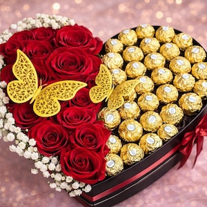 Pode incluir: Uma caixa de presente em forma de coração dividida em duas seções. Um lado apresenta rosas vermelhas e decorações de borboletas amarelas, o outro é preenchido com chocolates dourados. Uma fita vermelha adorna a caixa.