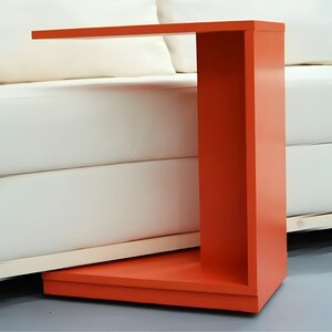 May include: A modern, orange side table with a cantilevered top. The table is a vibrant, solid color and has a rectangular base. The table is placed next to a white sofa.