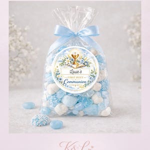 Puede incluir: Una bolsa transparente llena de caramelos azules y blancos, atada con una cinta azul claro. La bolsa presenta una pegatina redonda con el texto "Louie's First Holy Communion" y detalles florales. La imagen también muestra el texto "K&L Sweet Moments".