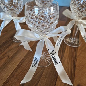 May include: Three clear crystal wine glasses with a diamond-cut pattern on the bowl and a starburst design on the base. Each glass is tied with a white satin ribbon featuring black printed text that reads BRIDE, Niamh, and BRIDESMAID.