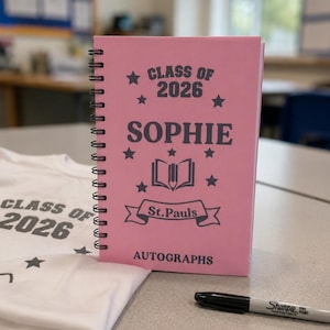 May include: A pink spiral-bound notebook with black text reading CLASS OF 2026, SOPHIE, St.Pauls, and AUTOGRAPHS, featuring star icons and an open book graphic. Beside it lies a white t-shirt with matching text and a black Sharpie fine point marker.