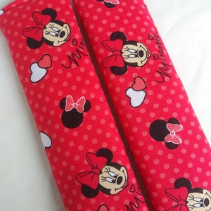 May include: Two red seatbelt covers with a polka dot pattern and Minnie Mouse illustrations. The covers feature Minnie Mouse's face, bows, and the word "Minnie" in cursive. White and red hearts are also present.