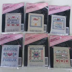 May include: Six Banar Designs Scentimental Samplers cross stitch kits in individual plastic packaging. Each kit features a different counted cross stitch pattern, including geese, a house, an alphabet, and hearts, with text like Country at Heart.