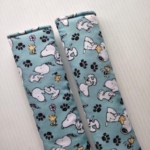 May include: Two light blue seatbelt covers with a repeating pattern of Snoopy, Woodstock, paw prints, and flowers. The covers are rectangular and padded, designed to fit over a seatbelt.