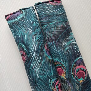 May include: Two fabric seat belt covers with a peacock feather design. The covers are primarily teal with accents of pink, purple, and white. The design features a detailed pattern of peacock feathers, creating a decorative and stylish accessory.