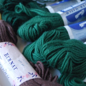 May include: A close-up of several skeins of yarn in various colors, including brown, green, and cream. The yarn is labeled "Bernat Foundation" and "Red Heart Super Saver".