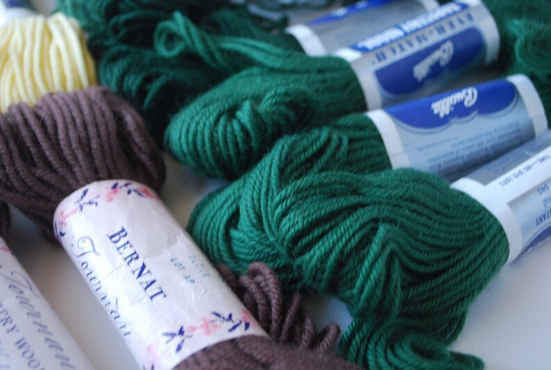 May include: A close-up of several skeins of yarn in various colors, including brown, green, and cream. The yarn is labeled "Bernat Foundation" and "Red Heart Super Saver".
