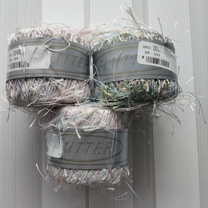 May include: Three skeins of yarn in shades of pink, green, and blue. The yarn is fluffy and has a shimmery texture. The skeins are labeled "Flutter" and have a SKU number of 001. DP: 084.