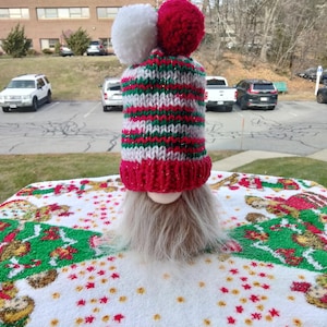 May include: A Christmas gnome with a knitted hat featuring red, green, and white stripes, topped with two fluffy pom-poms. The gnome has a long, gray beard and a round, pale face. The gnome is sitting on a white tablecloth with a Christmas-themed pattern.