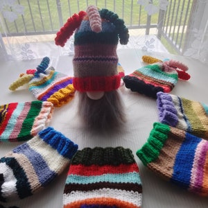 May include: A collection of colorful, hand-knitted gnome hats and beards. The hats feature a variety of striped patterns in shades of red, blue, green, and purple. The gnomes have long, gray beards and are arranged in a circle.