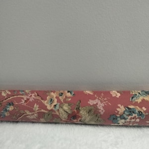 May include: A cylindrical door draft stopper with a floral pattern in shades of pink, blue, and cream. The fabric-covered stopper rests on a white, fluffy surface, offering a decorative and functional home accent.