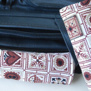 May include: Two fabric items with a repeating geometric pattern in red, brown, and beige. The pattern includes squares with heart, floral, and star-like designs. The items are placed near a black leather bag.