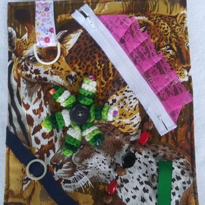 May include: A sensory activity board with a safari animal print background. Features include a white zipper, pink lace, a green and white crocheted flower, a navy blue ribbon, a green ribbon, and a beaded strand with red and brown beads.