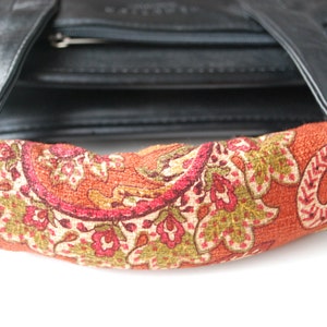 May include: A close-up of a black leather bag with a paisley patterned fabric handle. The handle is a bright orange color with green, red, and white paisley designs.