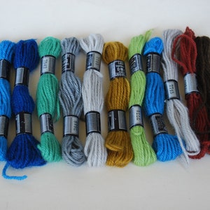 May include: A collection of embroidery floss in various colors, including shades of blue, green, gray, yellow, brown, and white. Each skein is labeled with a black and silver band. The floss is arranged on a white surface.