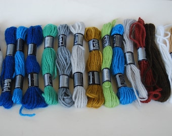 May include: A collection of embroidery floss in various colors, including shades of blue, green, gray, yellow, brown, and white. Each skein is labeled with a black and silver band. The floss is arranged on a white surface.