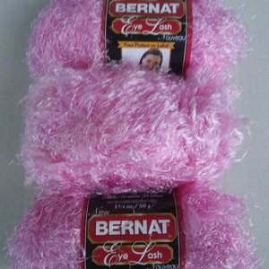 May include: Three skeins of pink Bernat Eye Lash yarn, a soft, fluffy yarn with a fuzzy texture. The yarn label reads "New Bernat Eye Lash Nouveau" and "Free Pattern on Label".