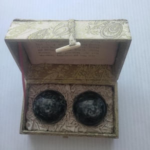 May include: A decorative box with a paisley pattern, containing two dark gray, marble-like spheres. The box is open, revealing a cream-colored interior and text that reads "Ball is a multi-ing device. It will become and doctor..."